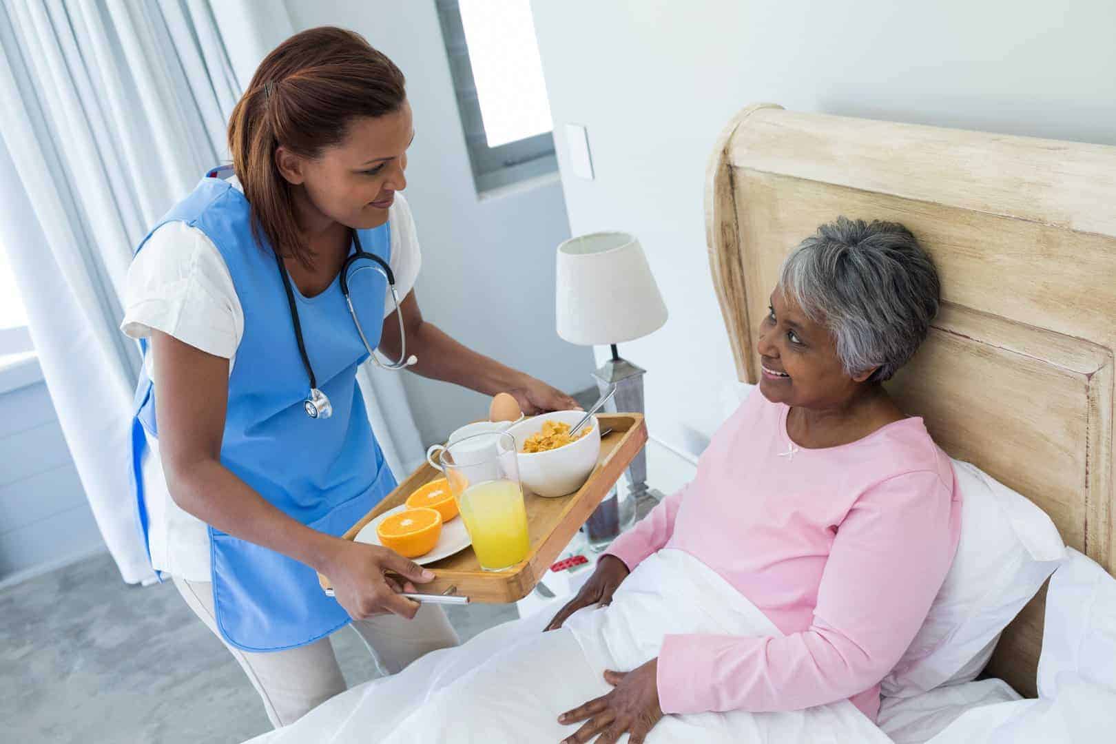 A caregiver in a blue uniform and apron serves breakfast to an older woman sitting up in bed, showcasing the compassionate services provided. The breakfast tray includes orange juice, toast, slices of orange, and a bowl of cereal.