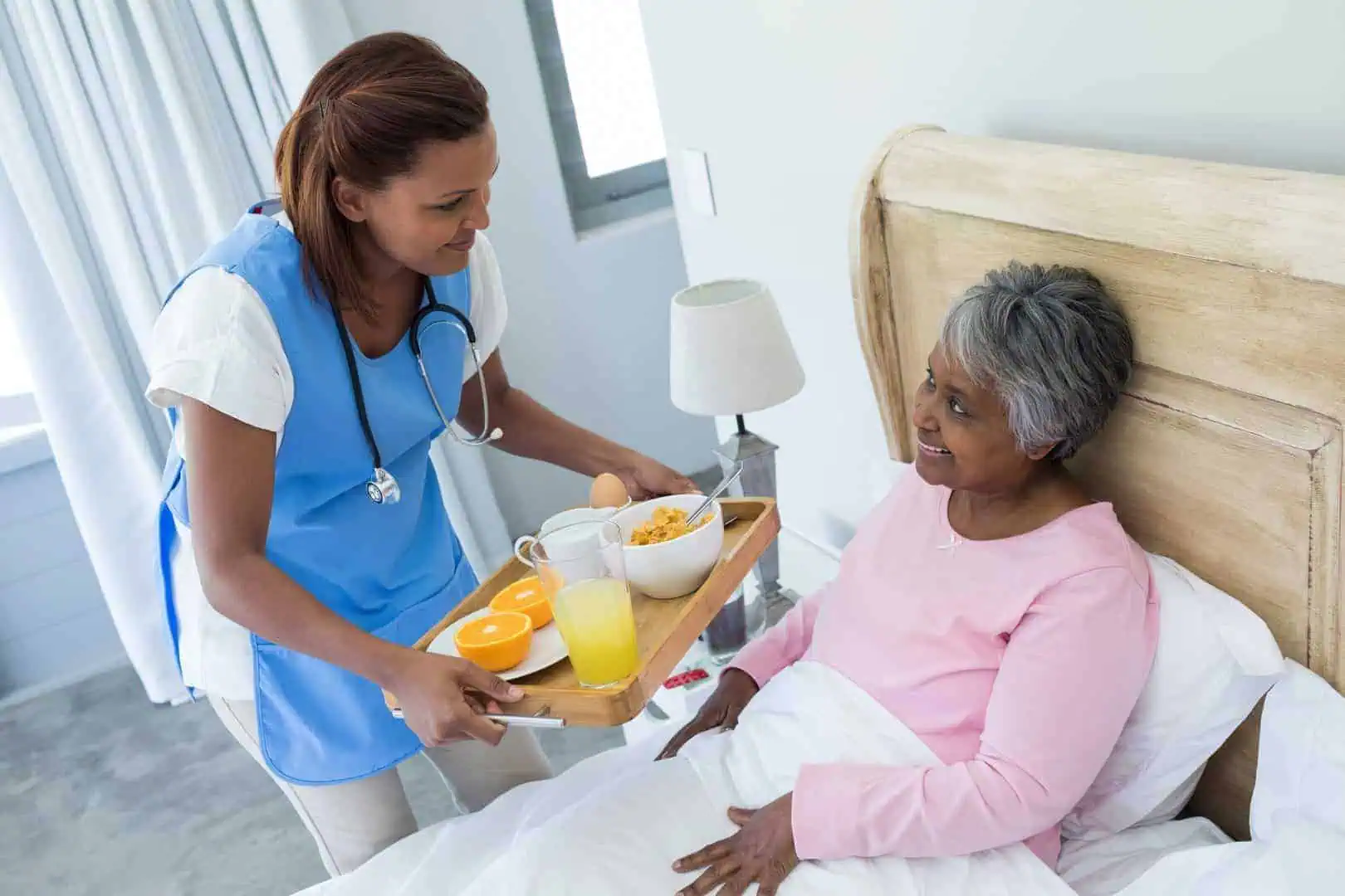 A caregiver in a blue uniform and apron serves breakfast to an older woman sitting up in bed, showcasing the compassionate services provided. The breakfast tray includes orange juice, toast, slices of orange, and a bowl of cereal.