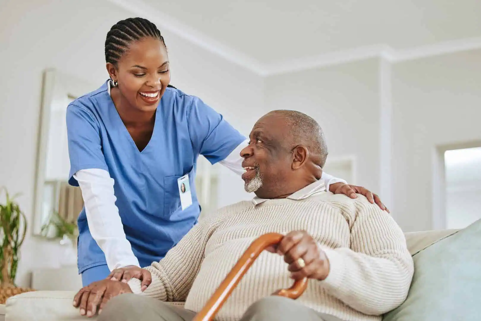 A nurse in blue scrubs smiles while providing physical therapy assistance to an elderly man with a cane sitting on a couch.