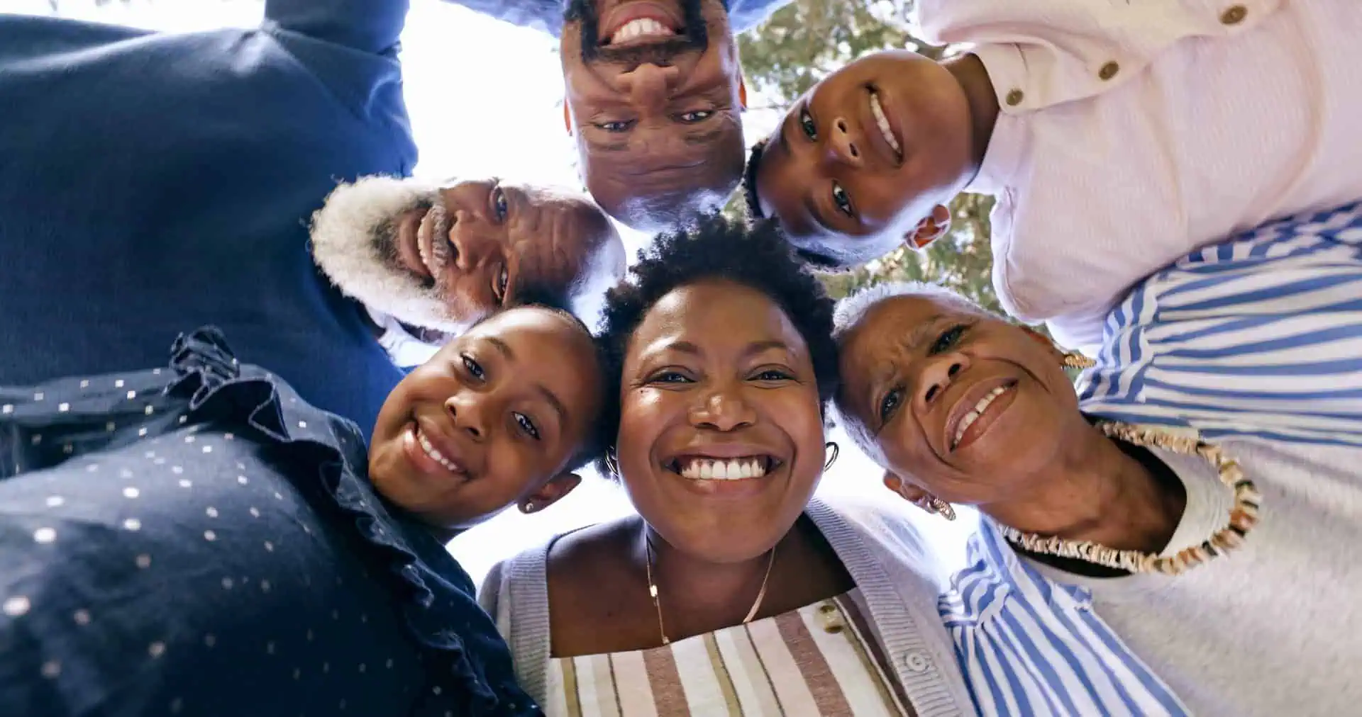 A group of six people, including two children and four adults, stands in a circle with heads touching, smiling and looking down at the camera, radiating joy and warmth as if sharing a moment of personal care.