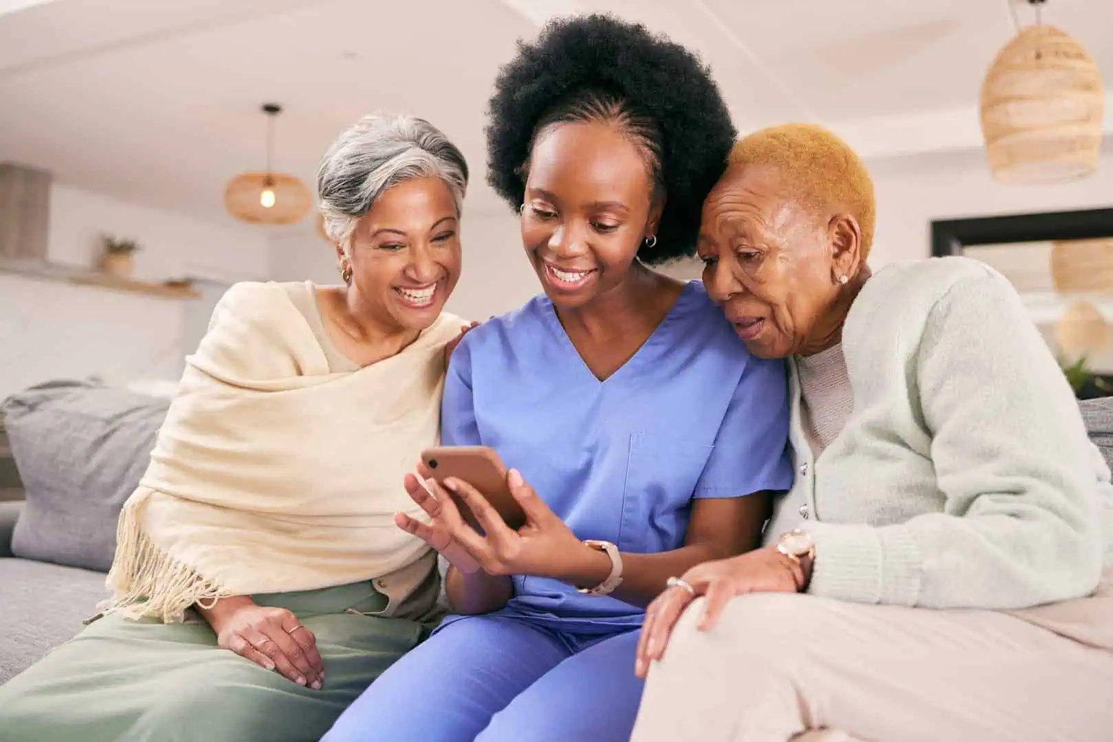 Three women sitting on a couch, looking at a smartphone together. One woman is in scrubs, suggesting she might be a healthcare worker. They appear engaged, smiling as they plan their day of running errands together.