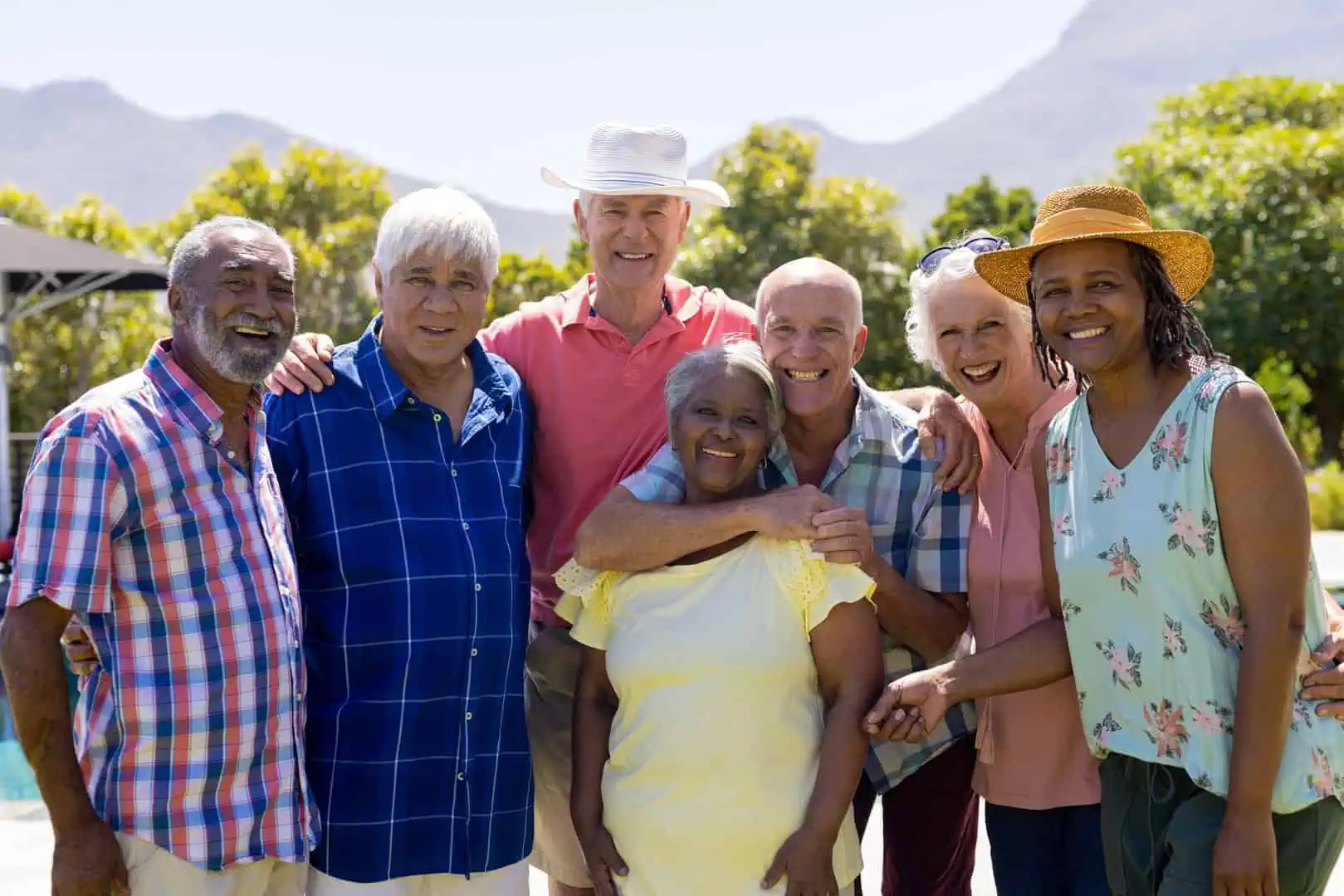 A group of six smiling senior friends embracing outdoors on a sunny day, enjoying personalized care, with mountains in the background.