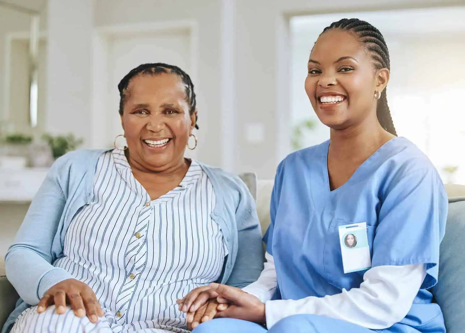 An elderly woman and a female nurse from Home Care Services in Toronto, Ontario, smiling at the camera, sitting together in a brightly lit living room.