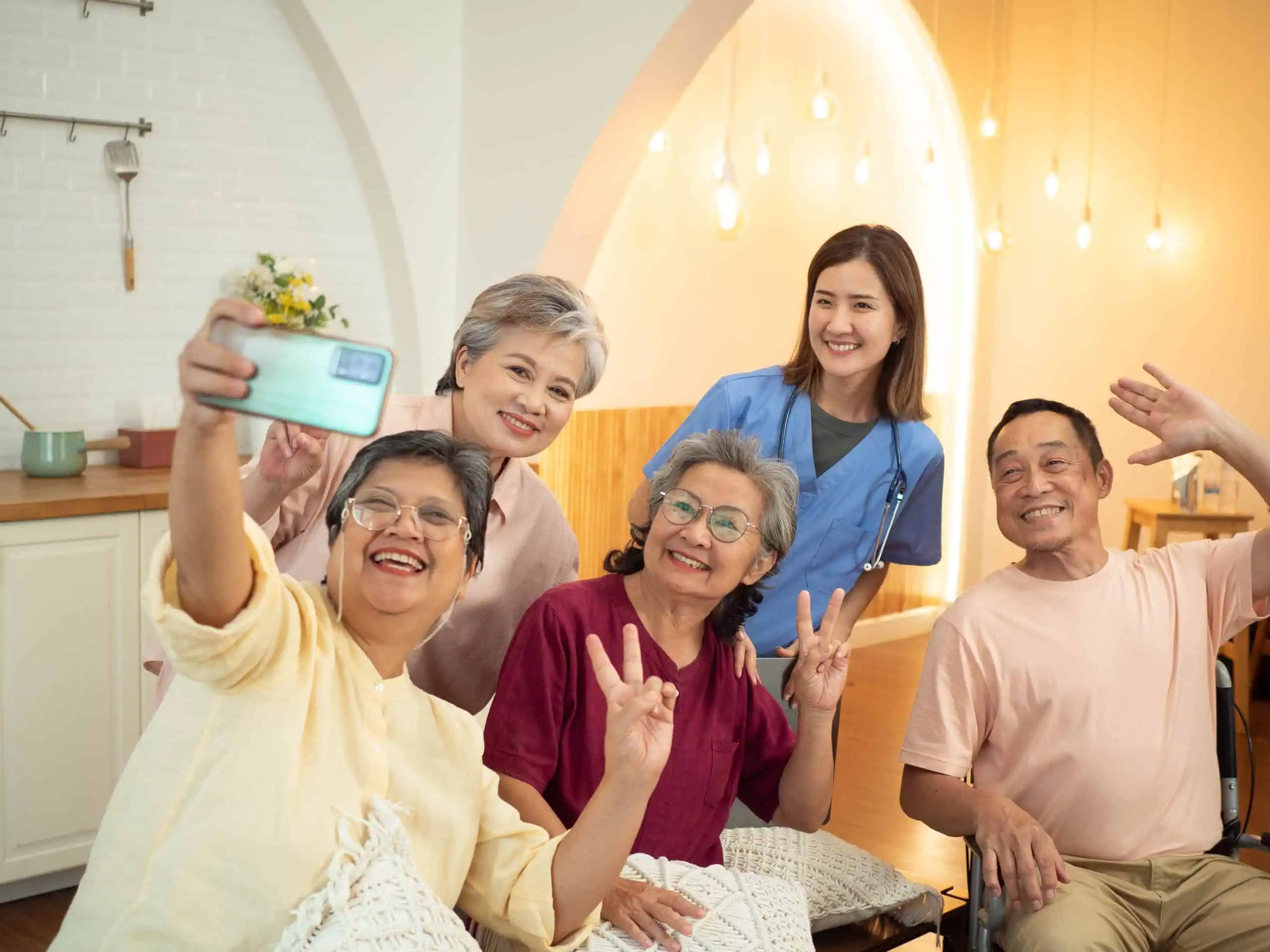 A group of five elderly individuals and one healthcare worker are sitting in a living room, smiling and making peace signs, capturing a moment of companionship with a cheerful selfie.