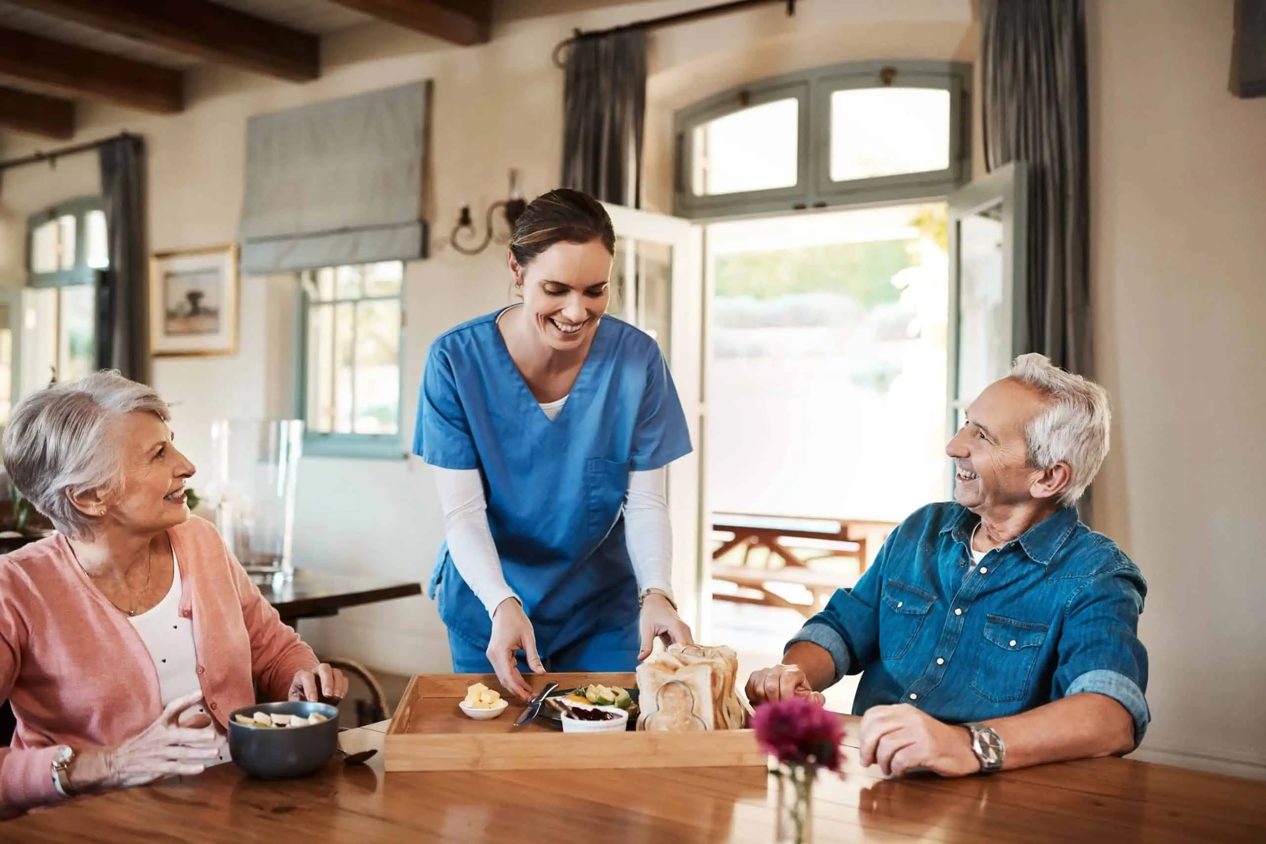 A caregiver in blue scrubs offers household assistance, serving meals to an elderly man and woman seated at a dining table in a well-lit room.