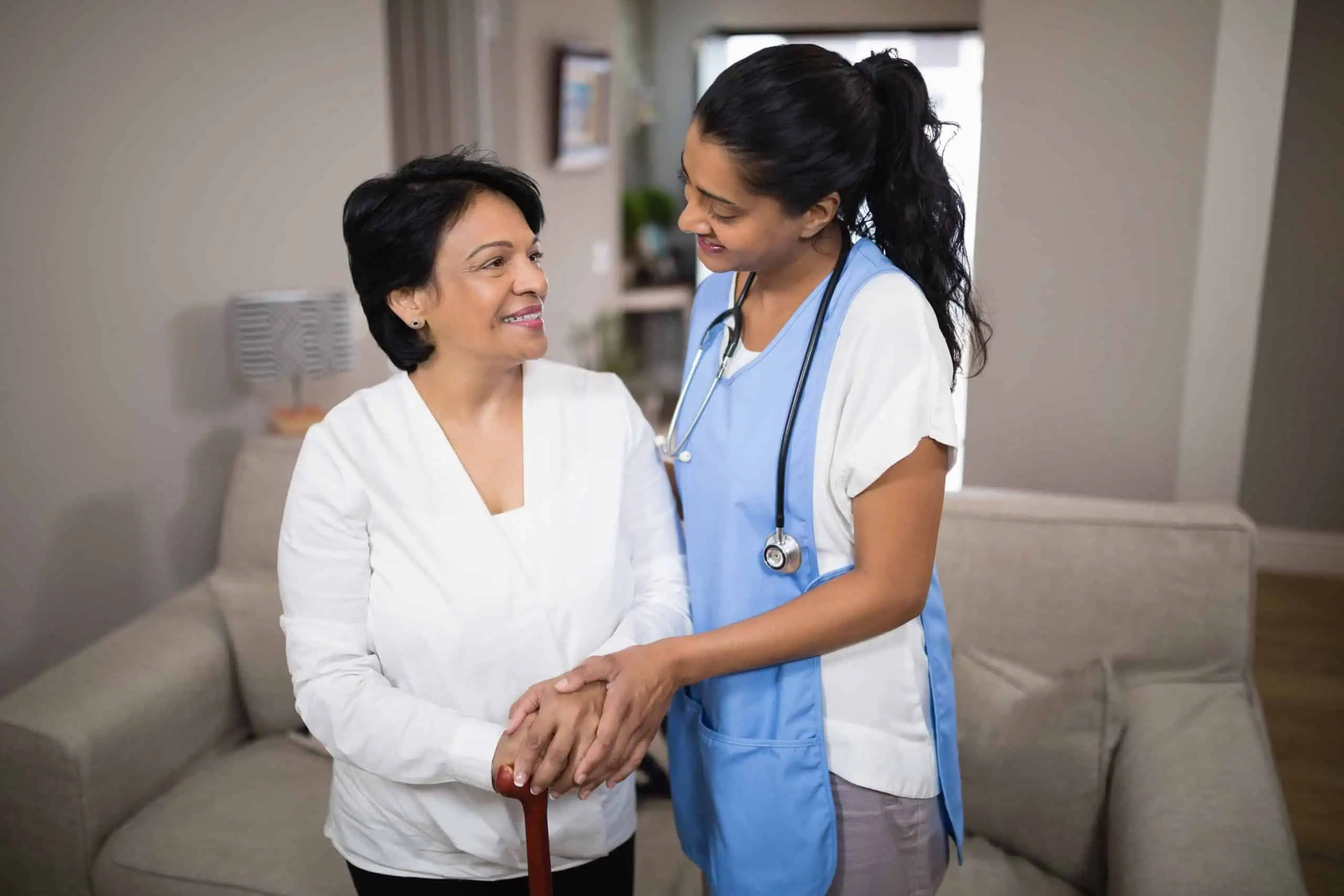 A nurse, wearing a stethoscope, stands and smiles while holding hands with an elderly woman who is holding a cane in a well-lit living room, emphasizing supervision and safety.