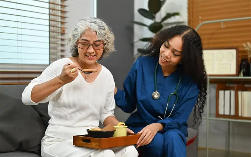 An elderly woman with gray hair sits on a couch, eating. A young healthcare worker in a blue uniform, providing household assistance, sits next to her, smiling and placing a hand on her shoulder. They appear to be in a living room.