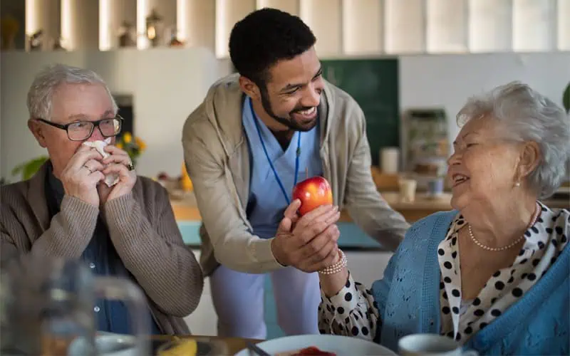 A caregiver in blue scrubs offers an apple to an elderly woman, exemplifying companionship, while an elderly man blows his nose at a table in a well-lit room.