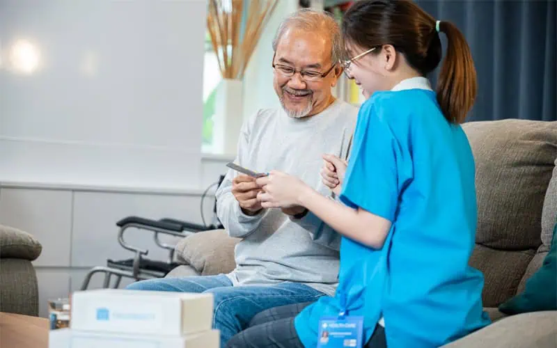 An elderly man sits on a couch smiling while a nurse in blue scrubs, who has been running errands, shows him something on a clipboard. A folded wheelchair is visible in the background.